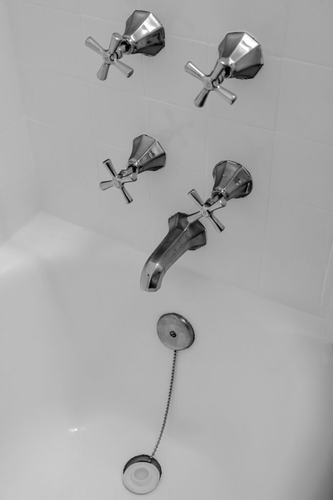Minimalist black and white image of a chrome bathtub faucet with cross handle knobs and chain plug.